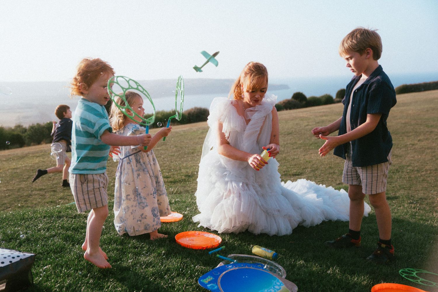 Bride in a flowing white tulle gown plays on a grassy hillside with children blowing bubbles and holding a toy airplane.