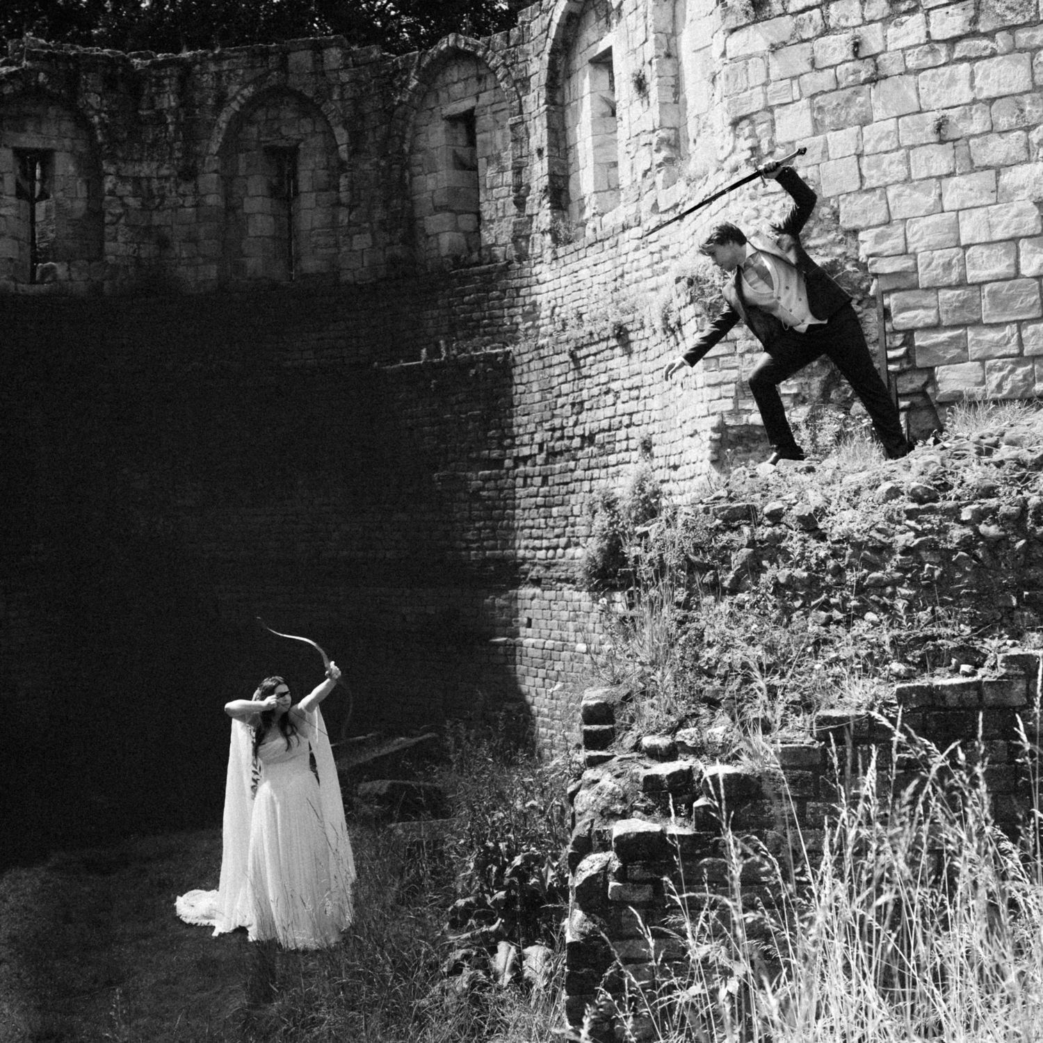 Black and white photo of a bride and groom near a rustic stone wall in a dramatic pose.
