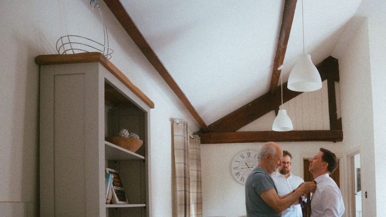 Two men adjusting a tie in a rustic room with wooden ceiling beams and a gray cabinet.