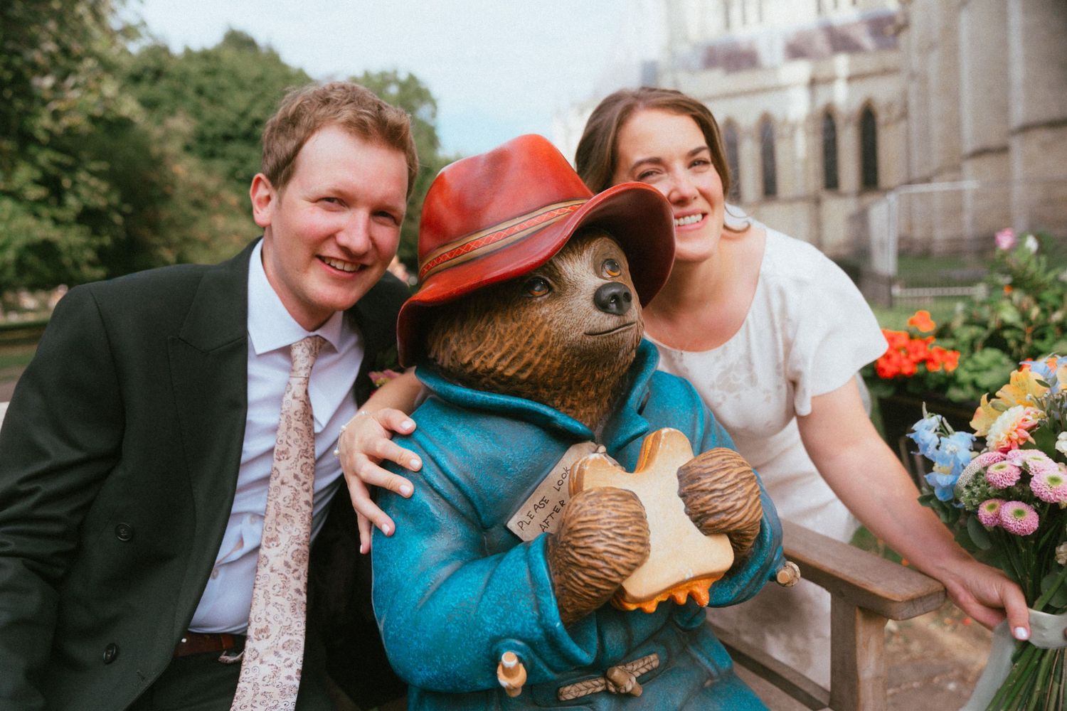 Bride and groom posing with a Paddington Bear statue on their wedding day outdoors.