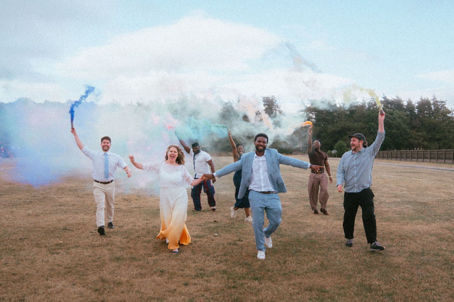 Wedding party running joyfully in a field holding colorful smoke bombs during an outdoor celebration.