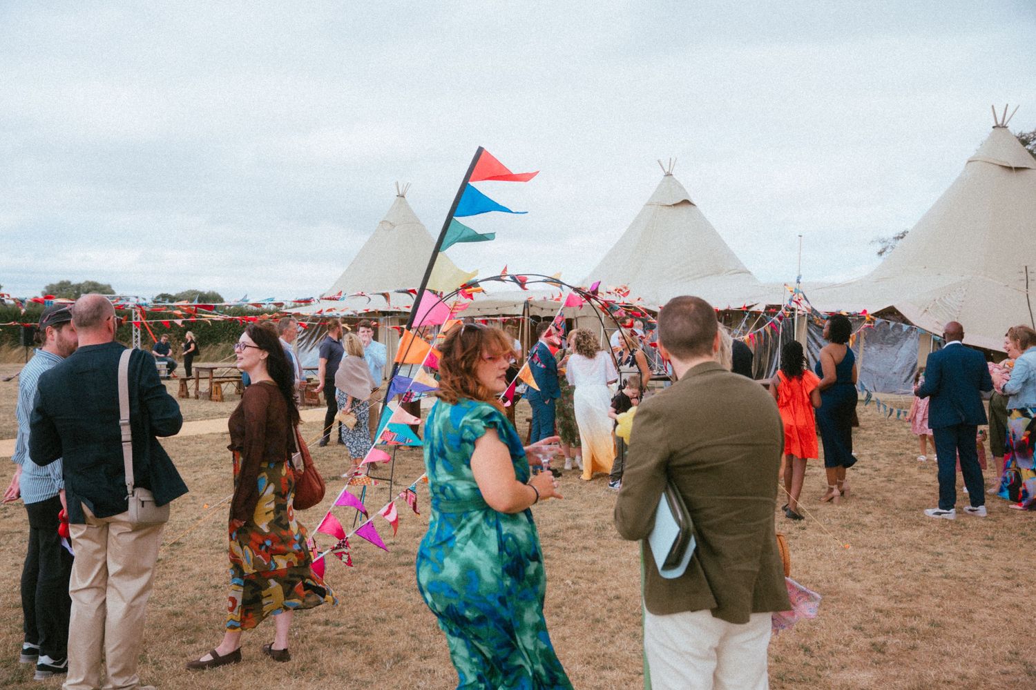 Guests mingle outside large tipi tents decorated with colorful flags at an outdoor festival event.