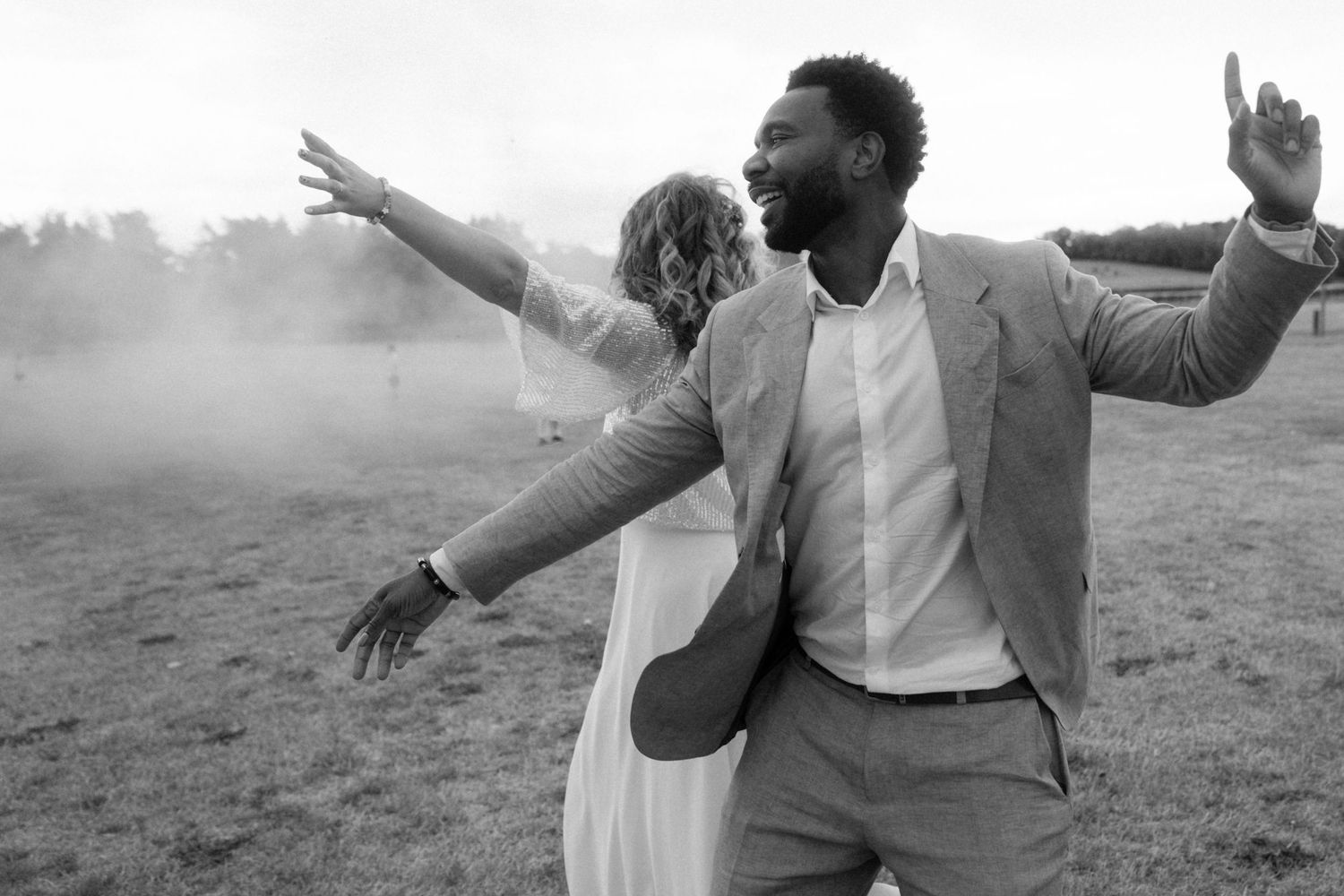 Black and white photo of a joyful couple celebrating outdoors with arms outstretched amid smoke at their wedding.