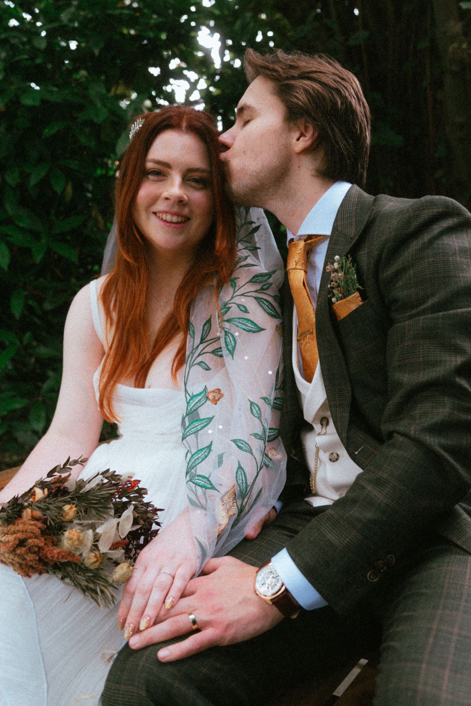 Groom in green tweed suit kisses bride with red hair wearing floral sleeve wedding dress while holding dried bouquet.