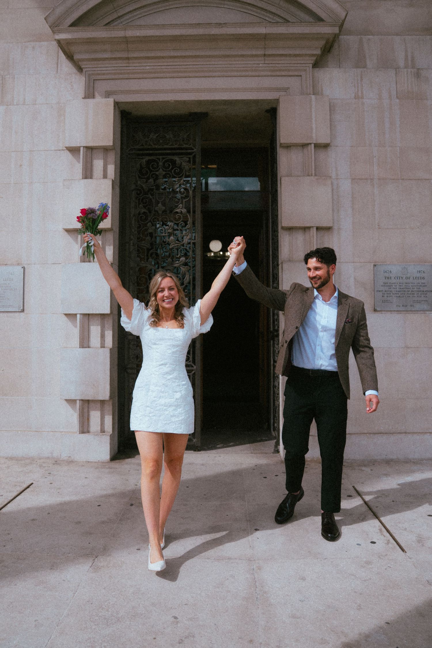 Joyful newlyweds exit a grand stone building, bride in white mini dress holding red roses, groom in brown suit, hands raised.