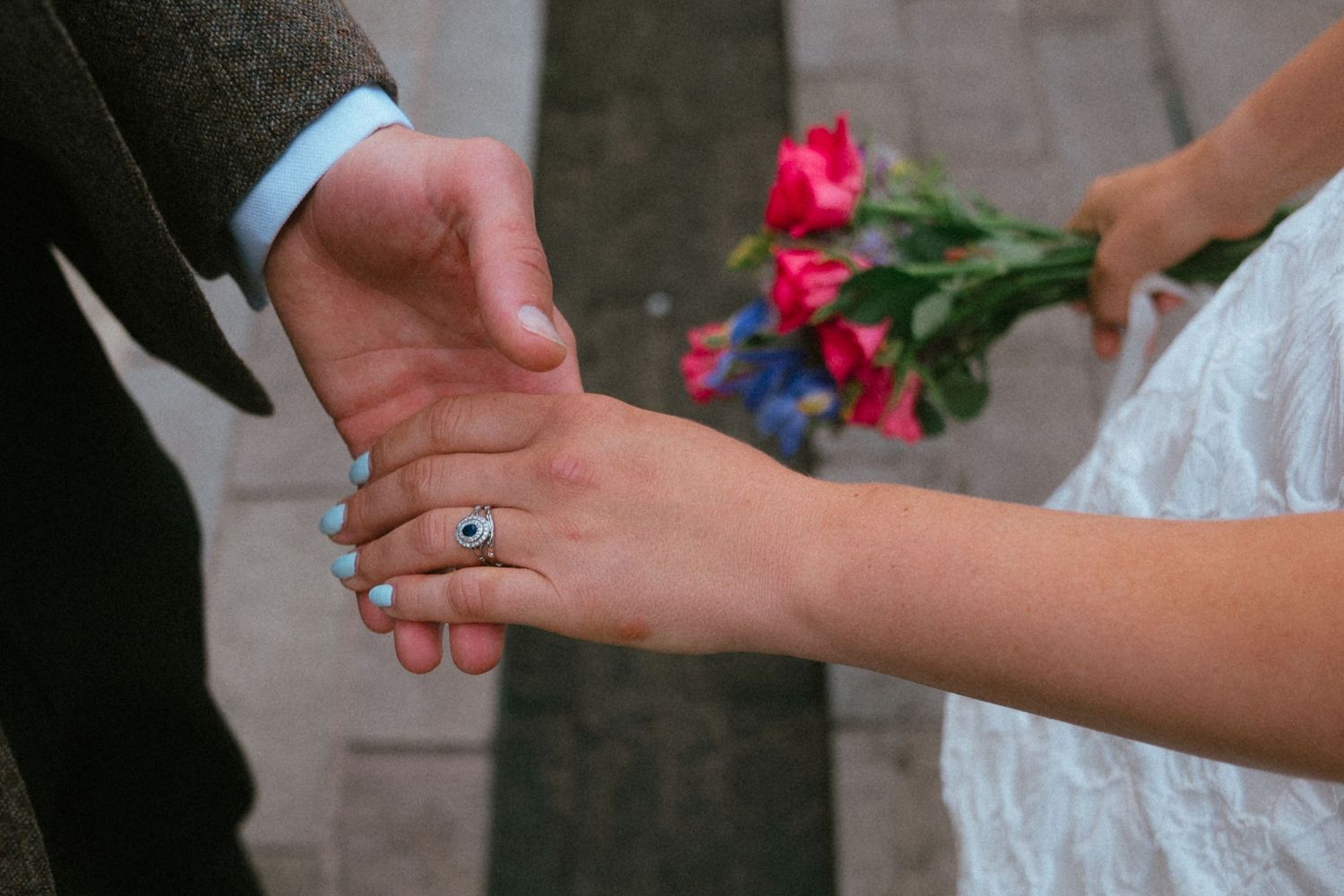 Close-up of bride and groom holding hands, showcasing an engagement ring and blue nail polish with a bouquet.