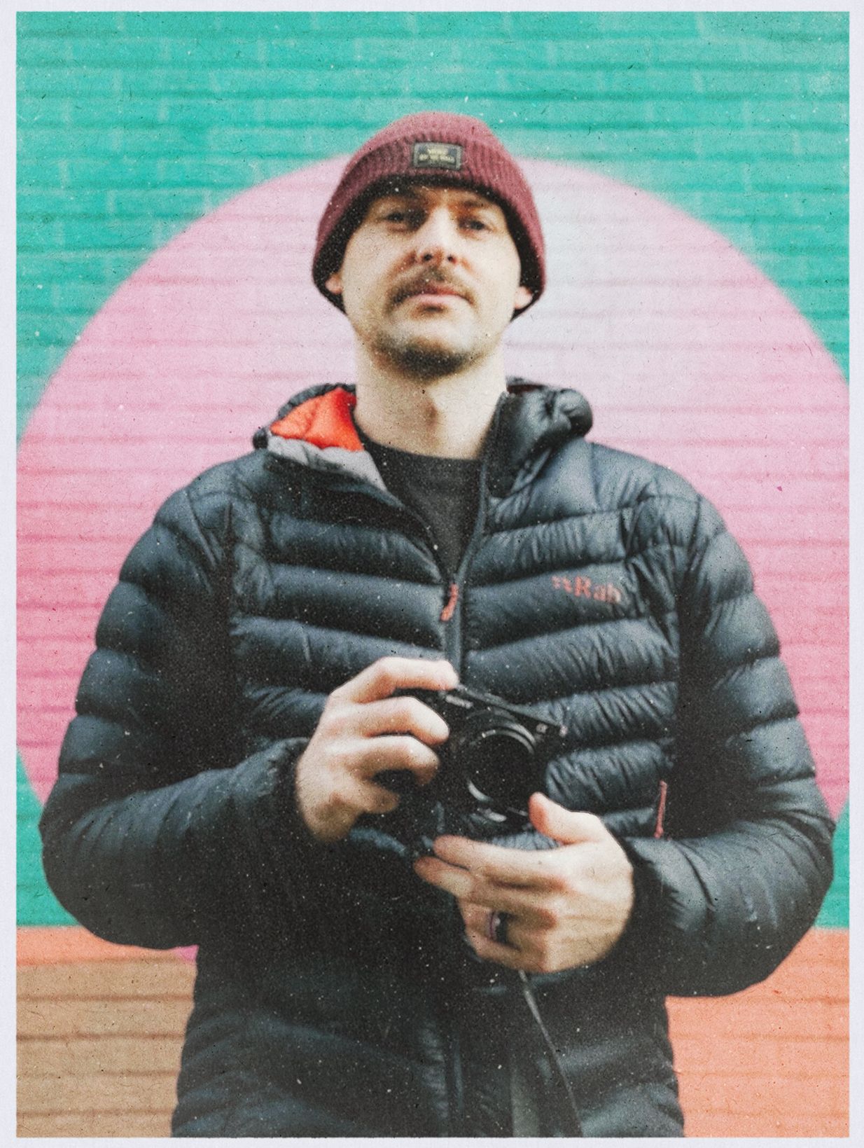 Male wedding photographer in puffer jacket holding camera against colorful pink and green geometric mural wall.