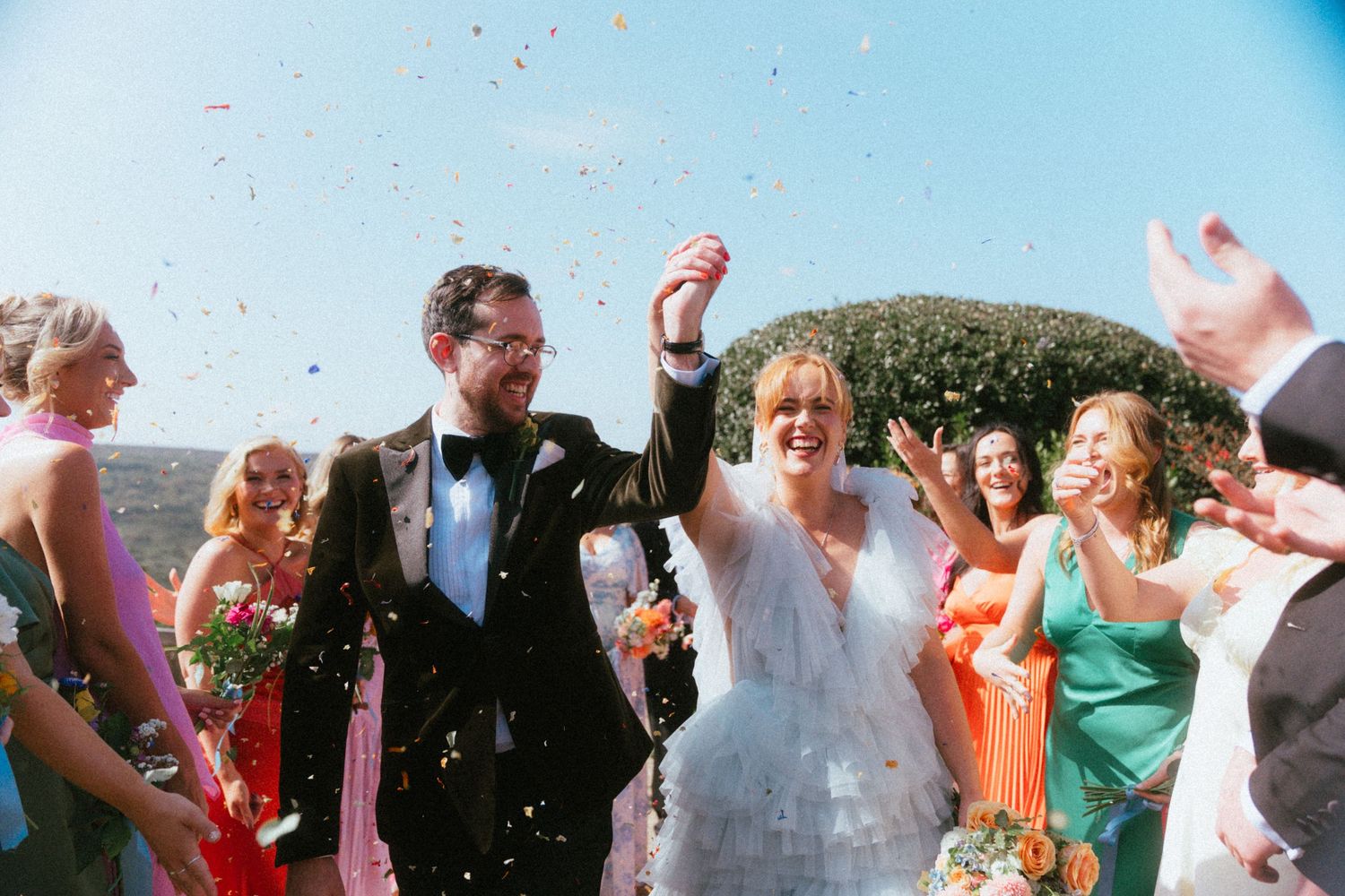 Bride in a white tulle gown and groom in a black tuxedo hold hands as wedding guests toss confetti around them outdoors.