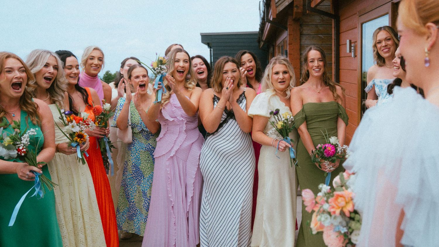 Group of smiling bridesmaids in colorful dresses holding bouquets of flowers and laughing together outdoors at a wedding.