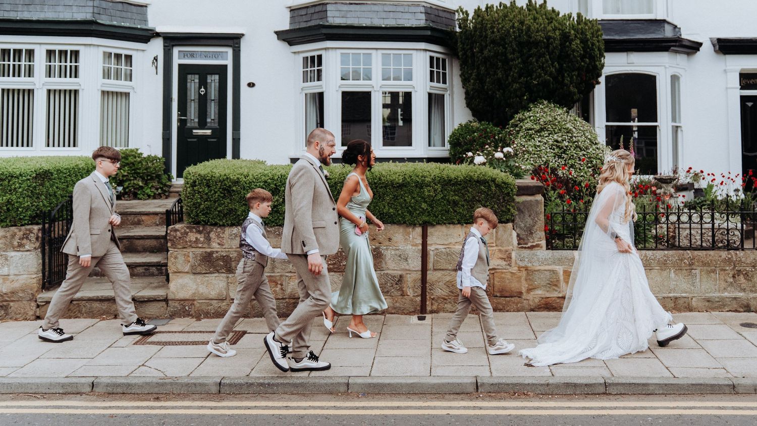 Wedding party walking in a line across a zebra crossing in an Abbey Road-inspired photo outside terraced houses.