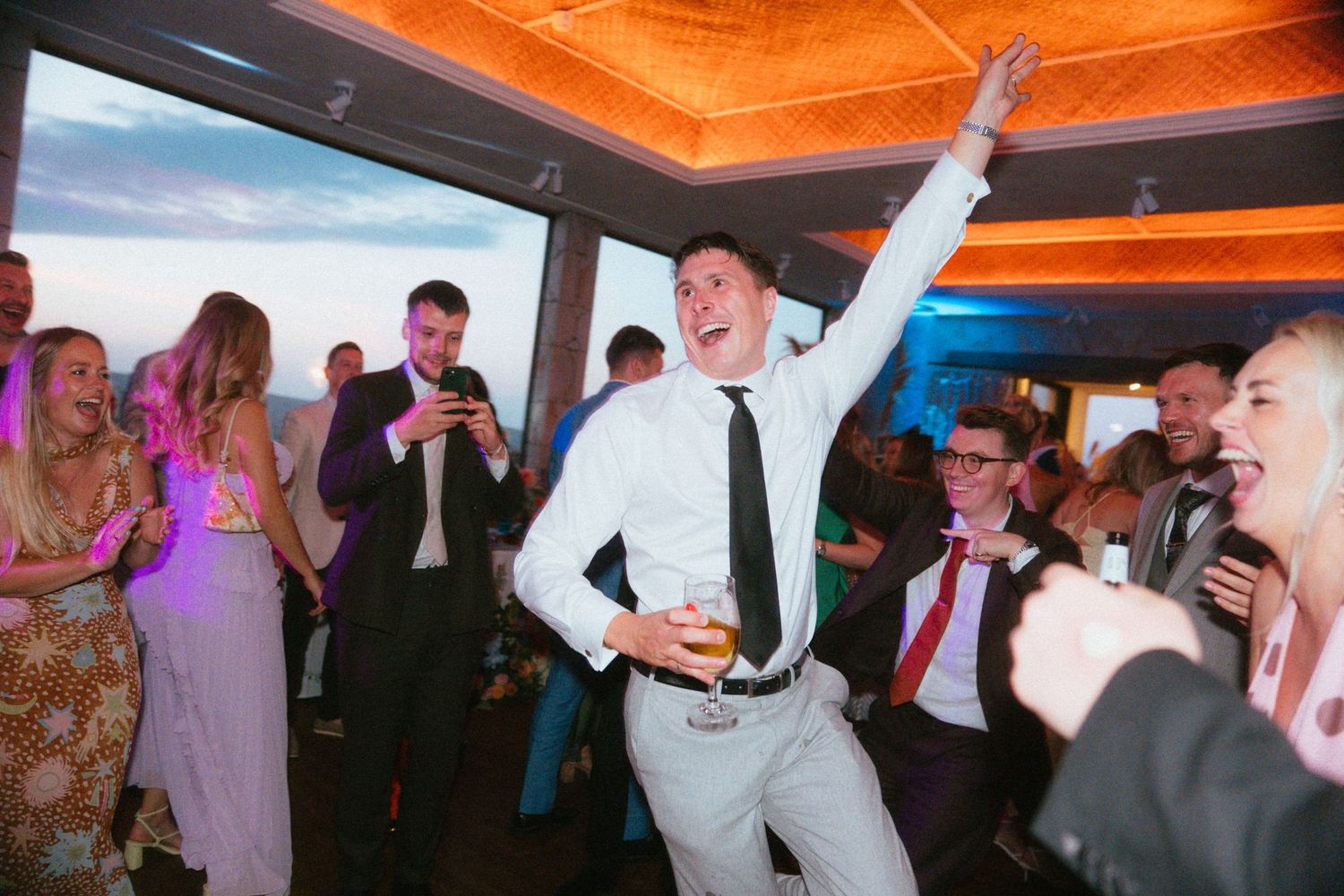 Smiling man in a white shirt and black tie raises one arm and holds a drink while dancing with guests at a lively indoor party.