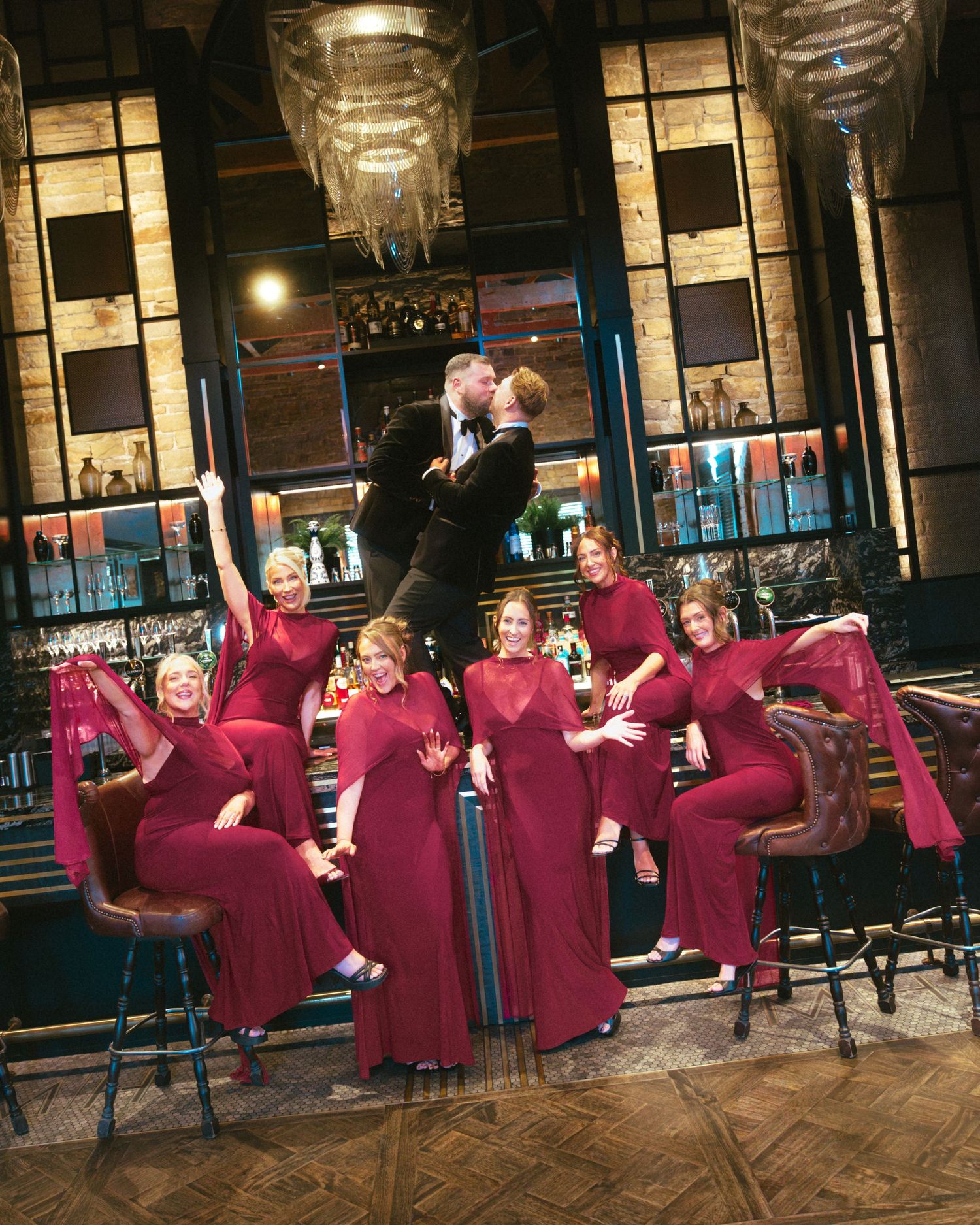 Bridal party in matching burgundy dresses poses with two grooms in dark suits at an upscale bar with chandeliers and leather stools.