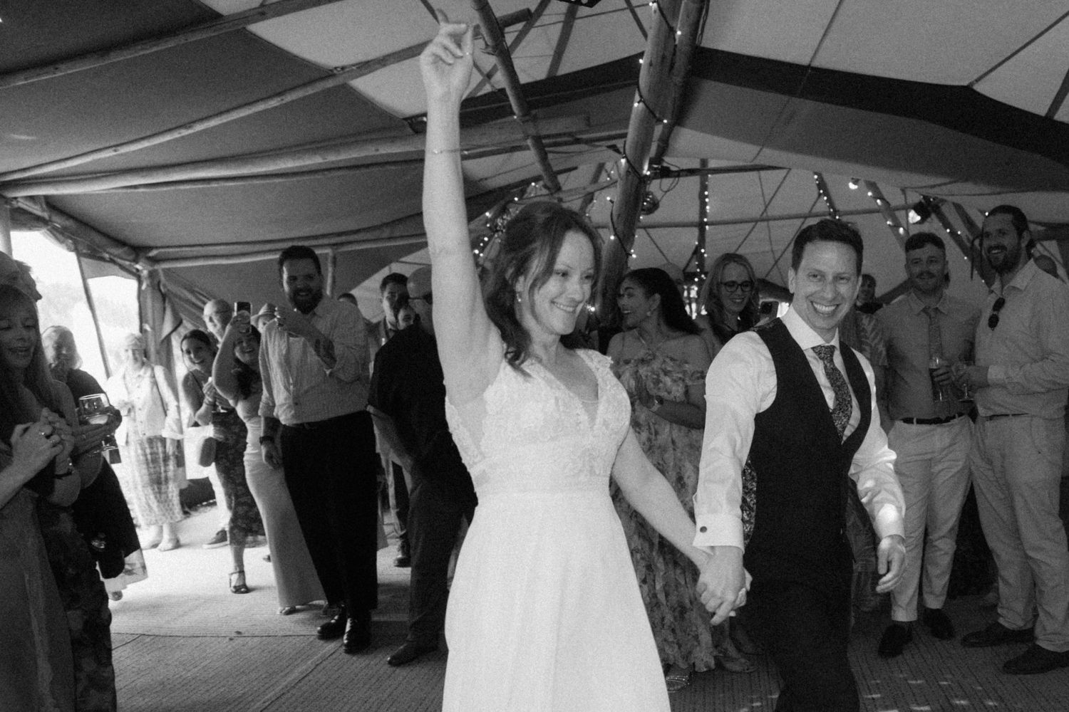 Black and white photo of jubilant bride and groom entering reception as guests celebrate around them.