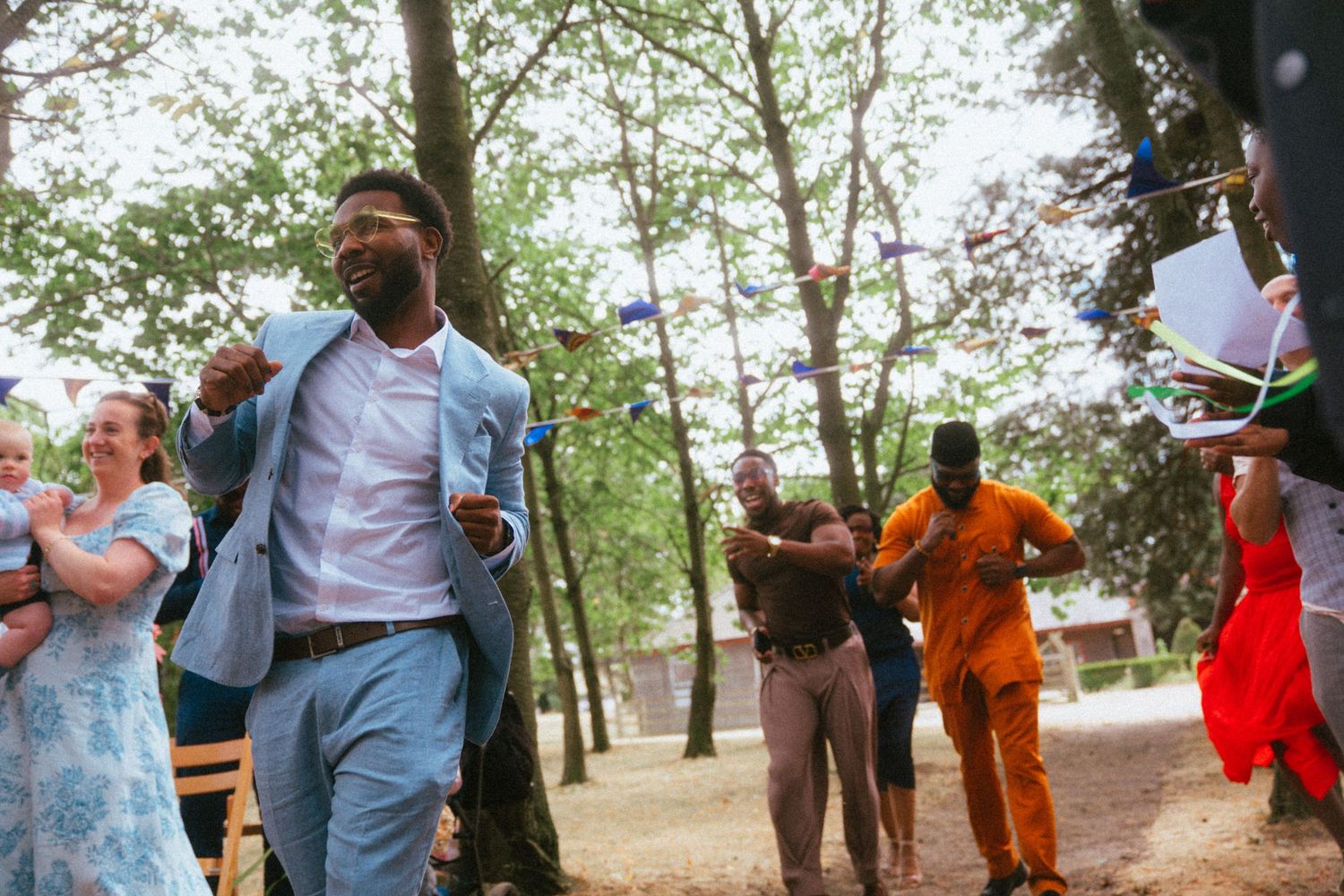A man in a light blue suit dances joyfully outdoors among trees at a festive celebration.