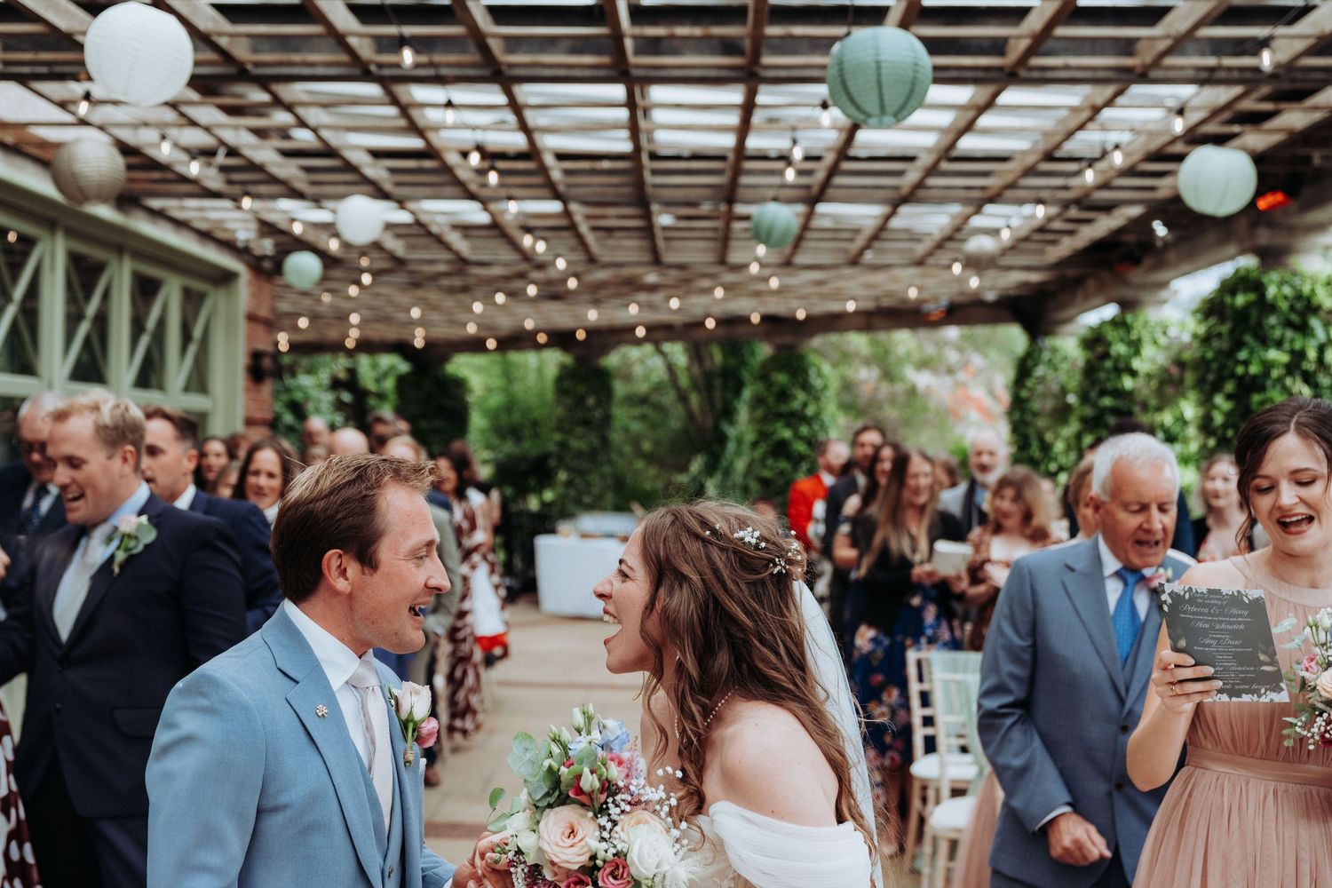 Happy bride and groom walking down aisle under pergola with string lights and paper lantern decorations.