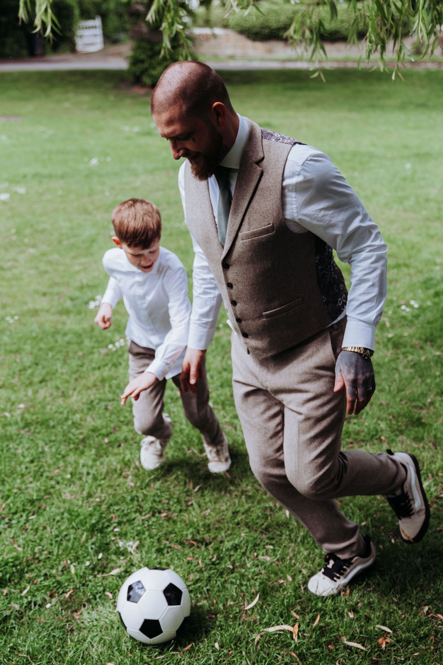 Tattooed groom in grey suit plays soccer with a young boy on a green lawn during wedding reception.