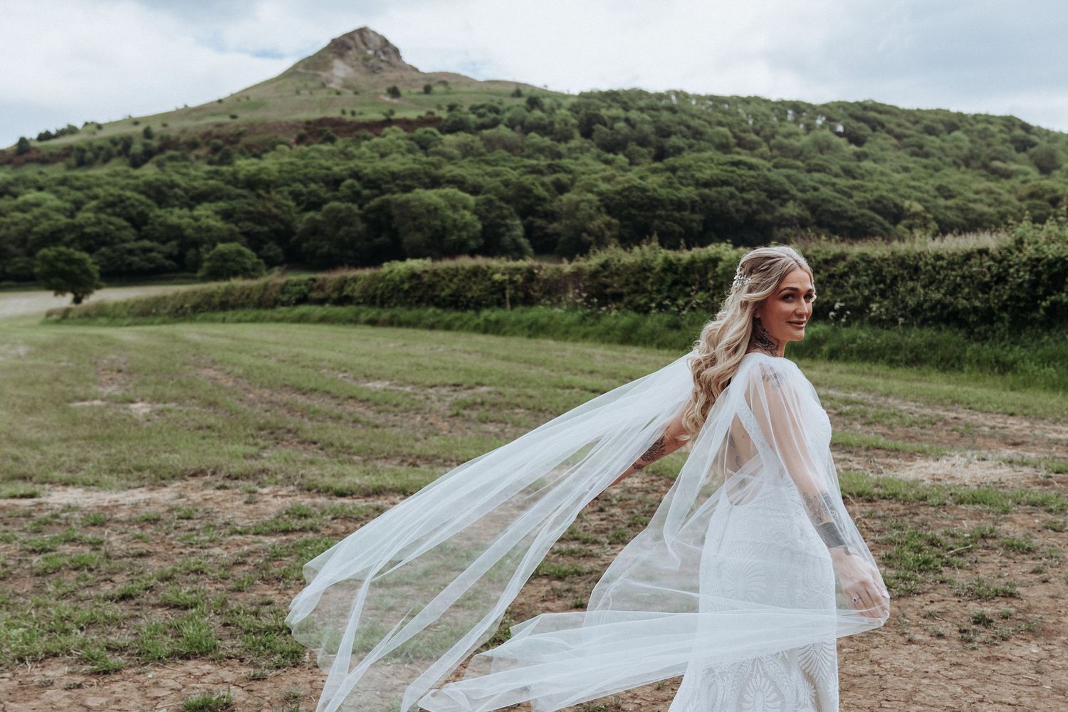 Bride in flowing white lace gown with dramatic veil billowing in wind against scenic hillside.