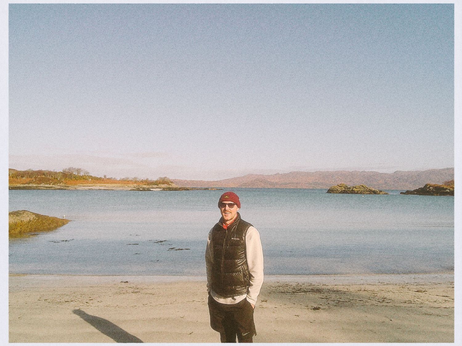 Man standing on a sandy beach with calm blue water and hills in the background on a clear day.