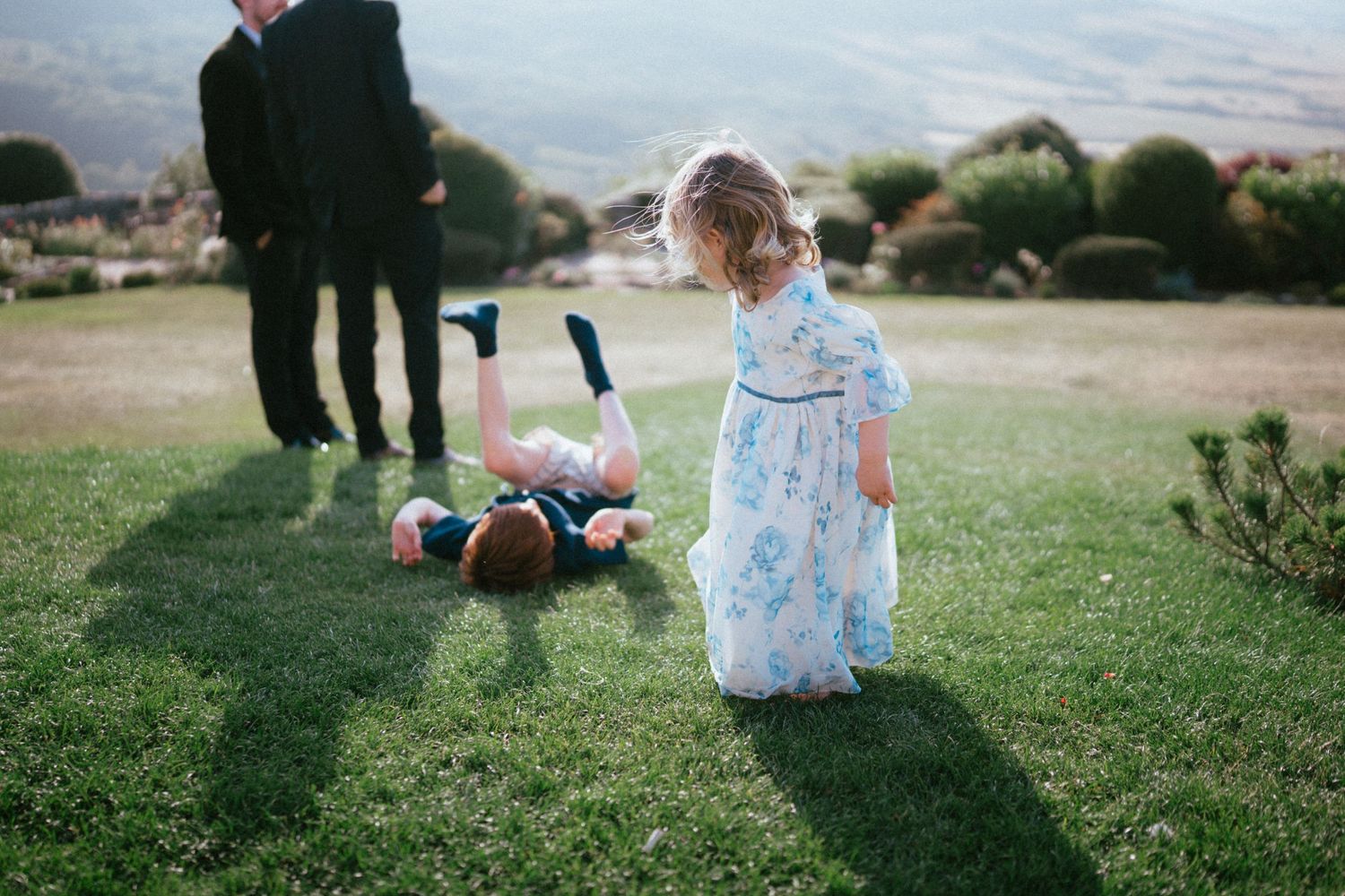 A little girl in a blue floral dress stands on a sunny lawn while a child lies on the grass near two men in dark suits.