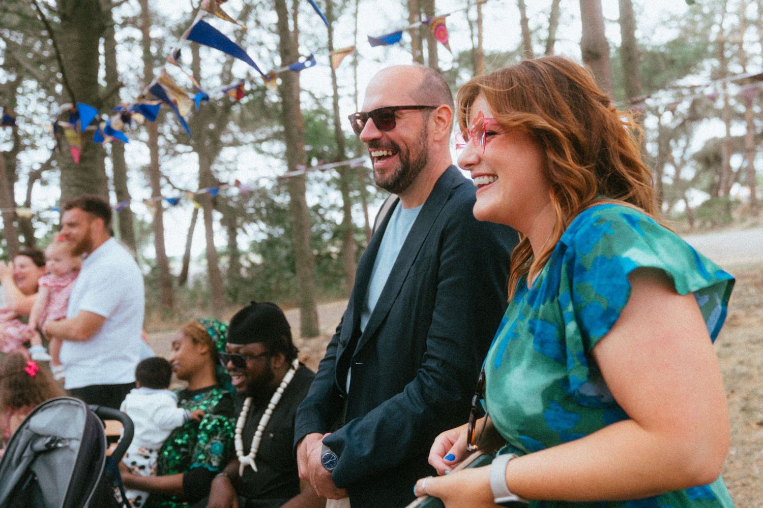 A smiling man and woman enjoy an outdoor celebration with colorful bunting and guests in a wooded setting.