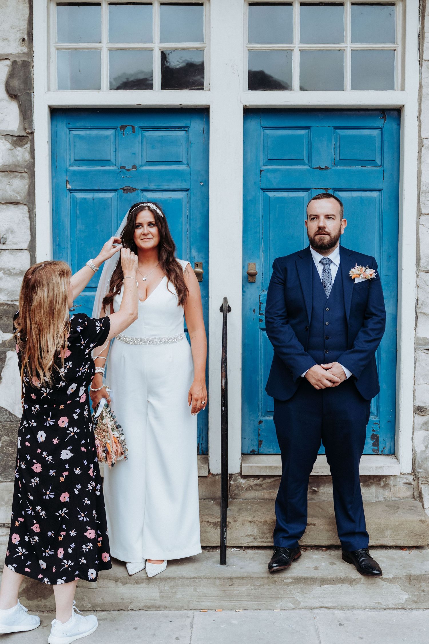 Bride and groom pose before blue doors while bridesmaid adjusts bride's hair on wedding day.