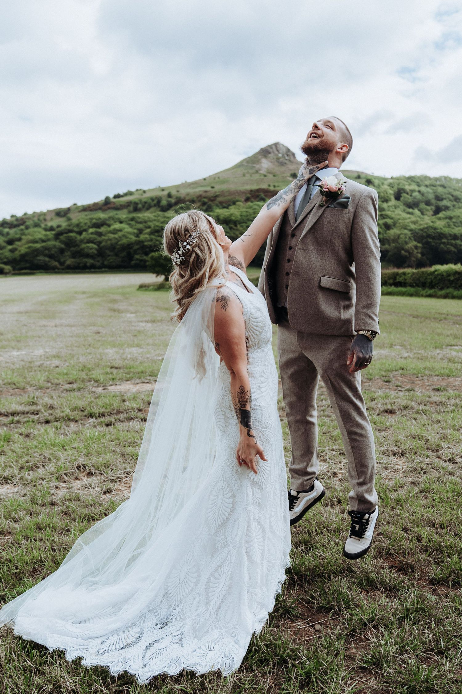 Bride adjusting tattooed grooms tie while he laughs in open green field with hill backdrop.