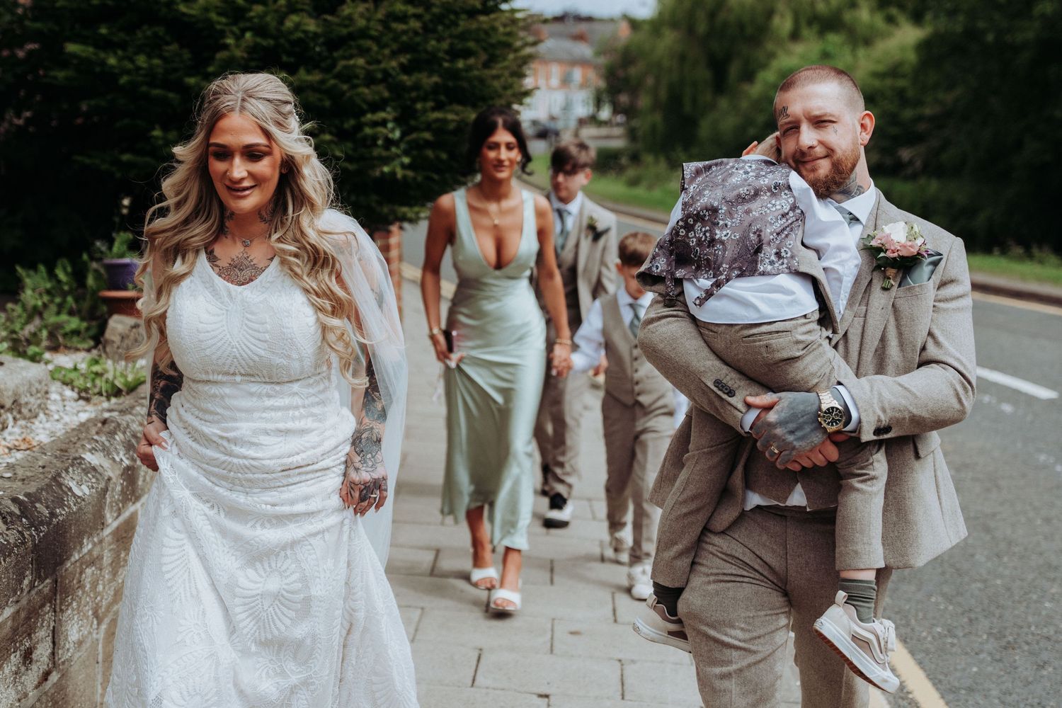 Tattooed bride in white gown walks with bridesmaids in sage green dresses as groom carries a child.