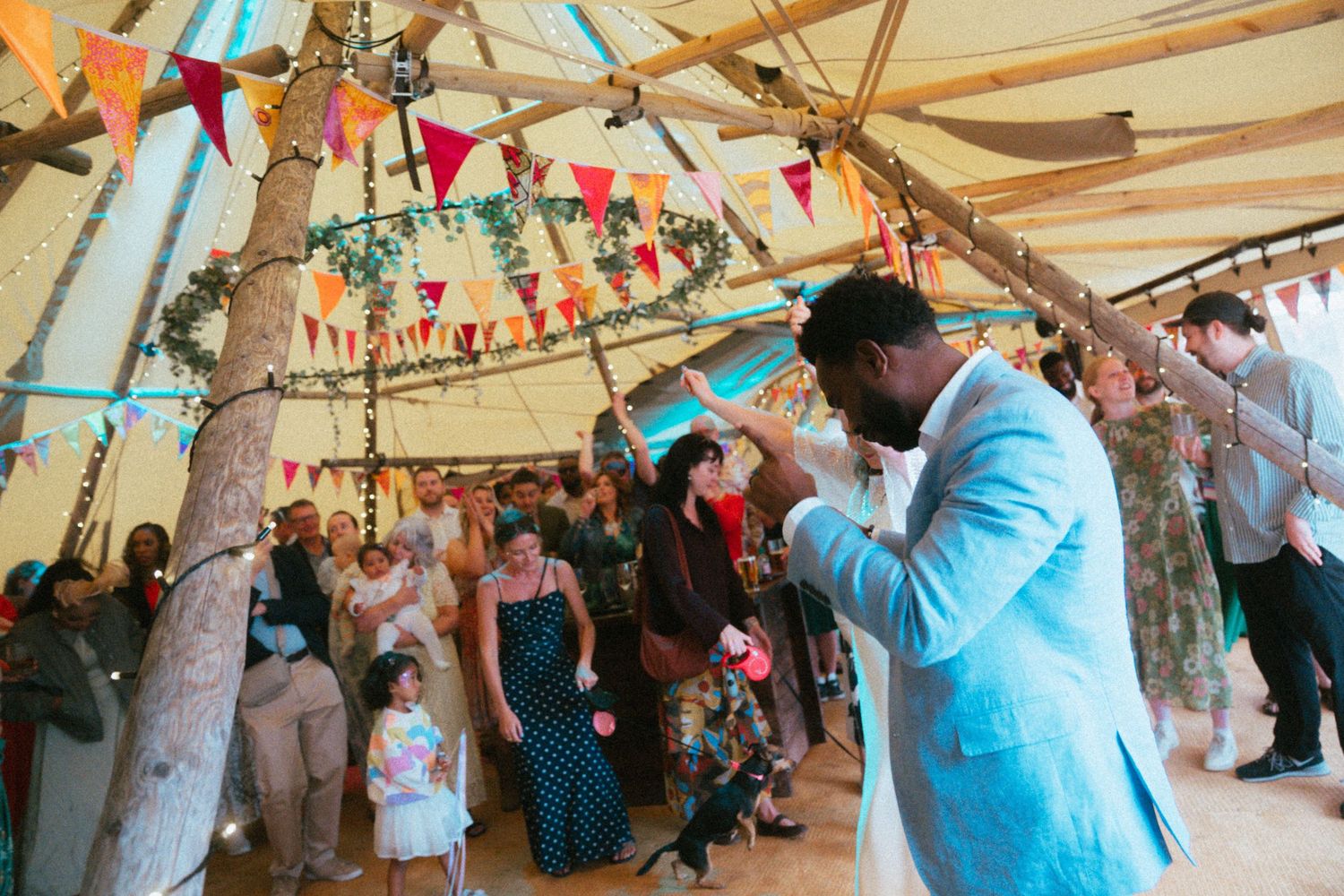 Groom in light blue suit celebrates under colorful bunting in a tipi wedding venue with guests in background.