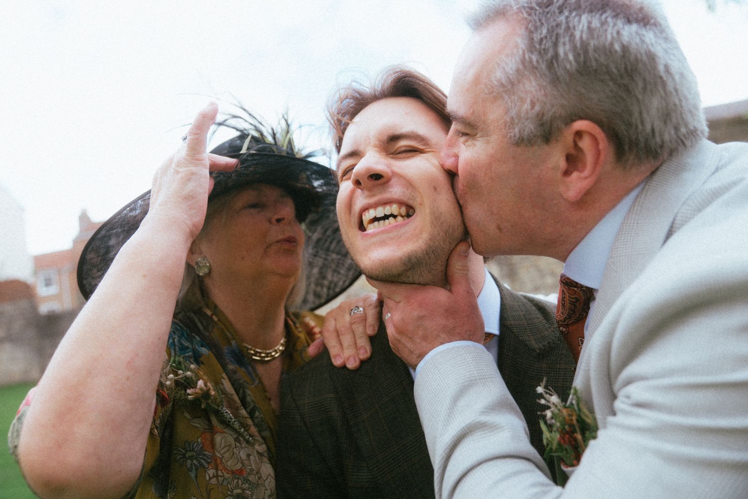 A joyful man receives kisses from an older couple at a wedding celebration outdoors.