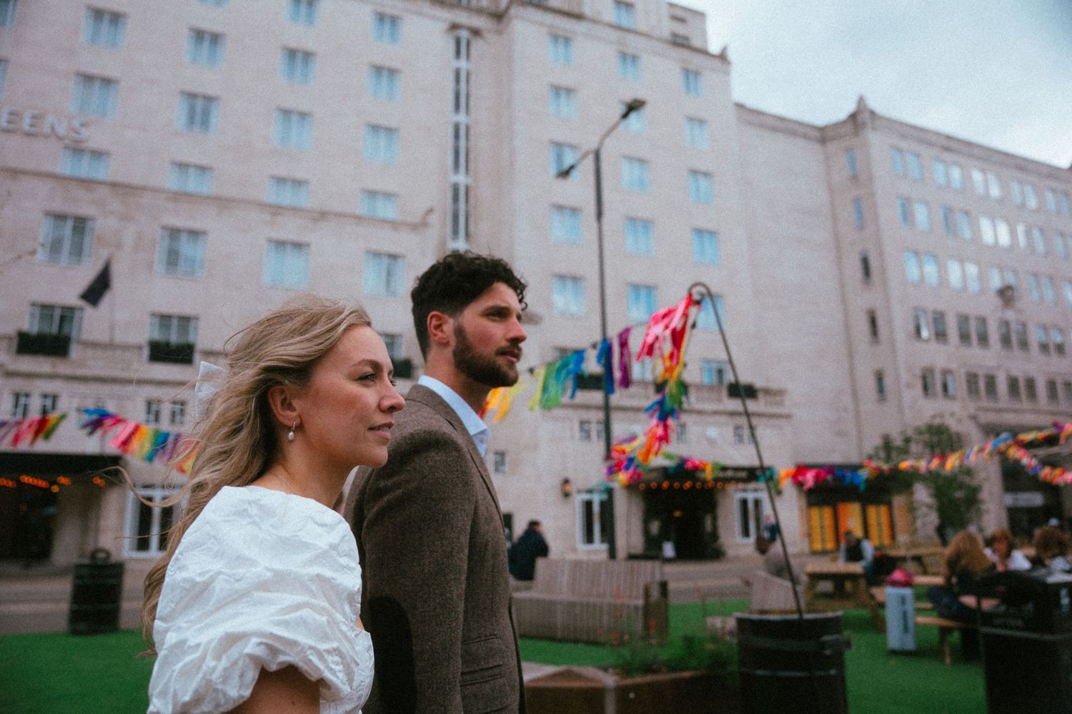 Elegant couple standing together outside grand hotel decorated with colorful bunting flags.