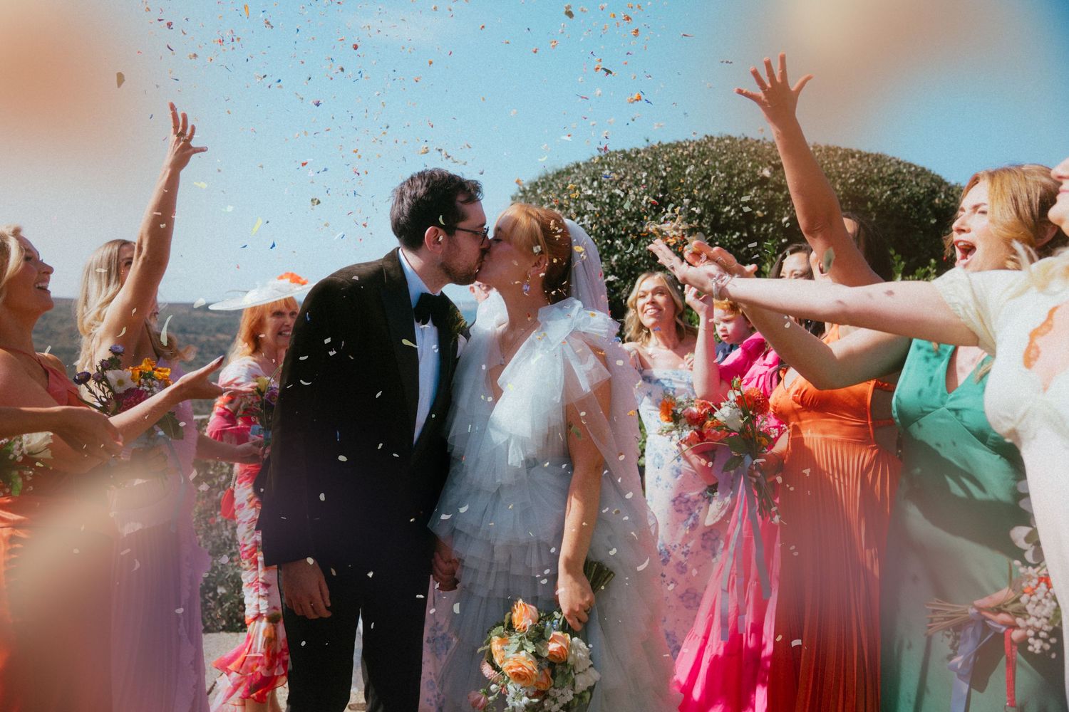 Bride and groom share a kiss as wedding guests in colorful dresses cheer and throw confetti in the air outdoors.