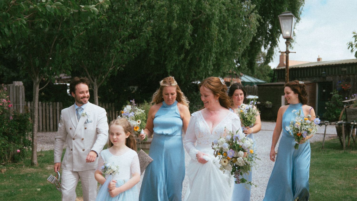 Bride in white gown walks with bridesmaids in blue dresses and flower girl on a sunny wedding day.