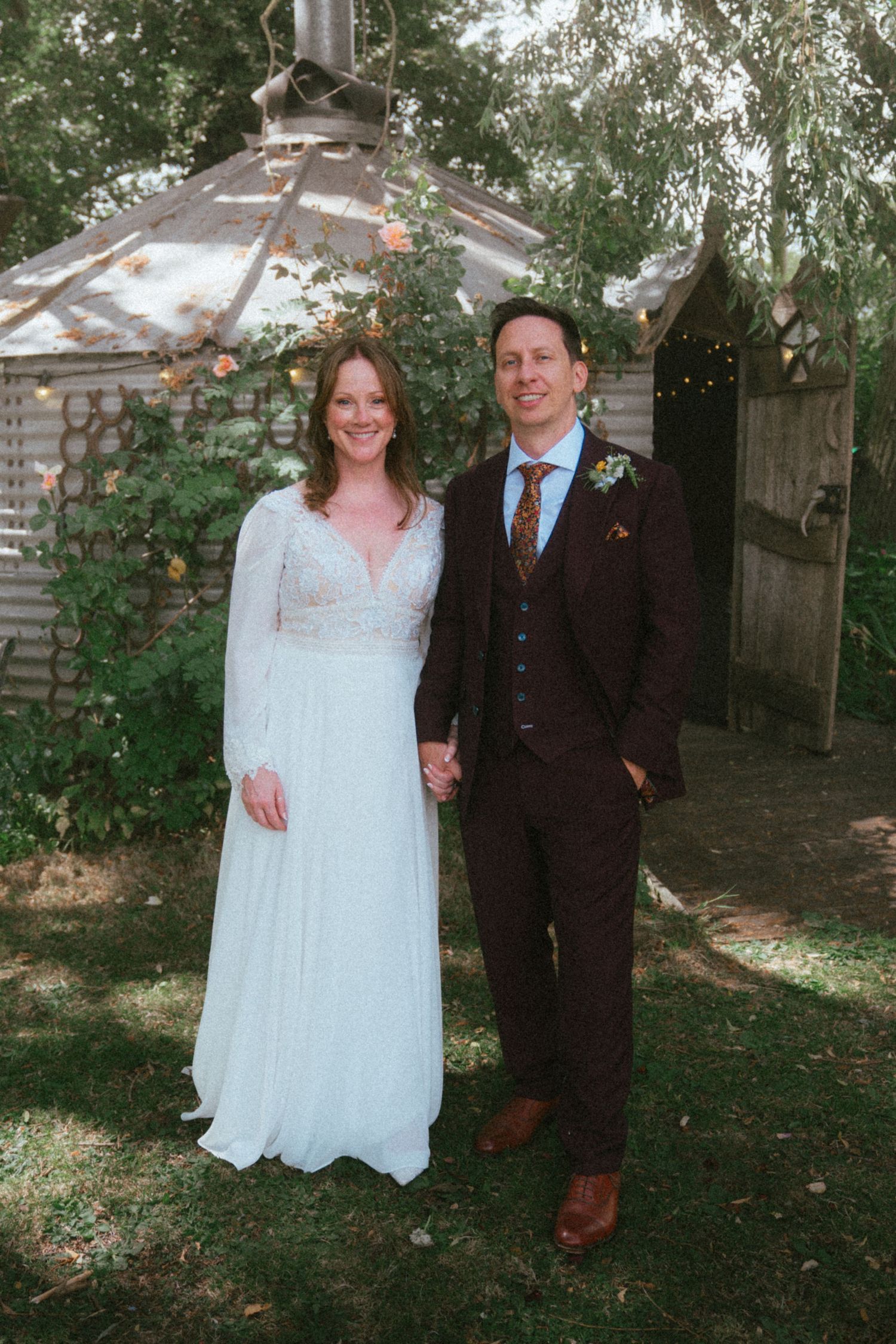 Smiling bride in lace long-sleeve gown and groom in brown suit pose together in garden setting.