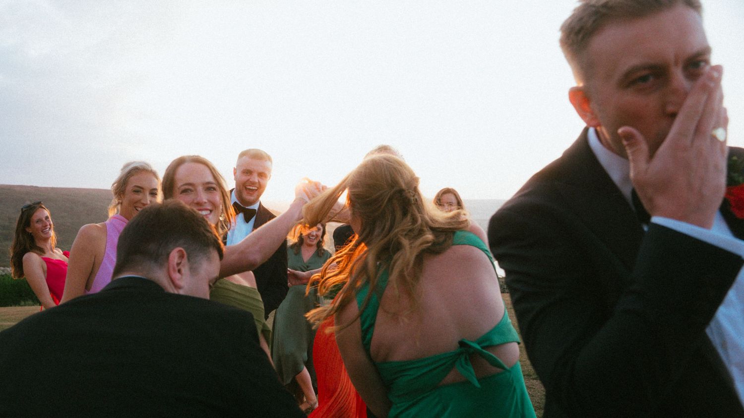 Wedding guests in formal attire laugh and embrace outdoors while a man in a dark suit covers his face with his hand.