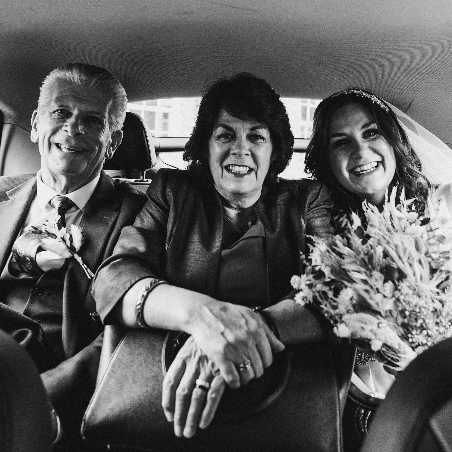 Black and white photo of smiling bride with parents in backseat of wedding car.
