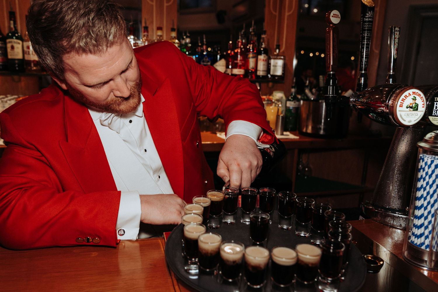 Man in red jacket carefully preparing a tray of Guinness shots at a wedding bar.