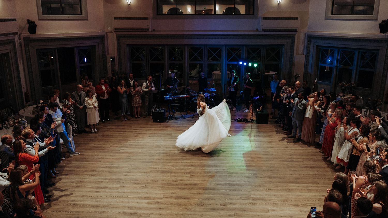 Bride twirling in a white gown during first dance on a wooden floor surrounded by guests.