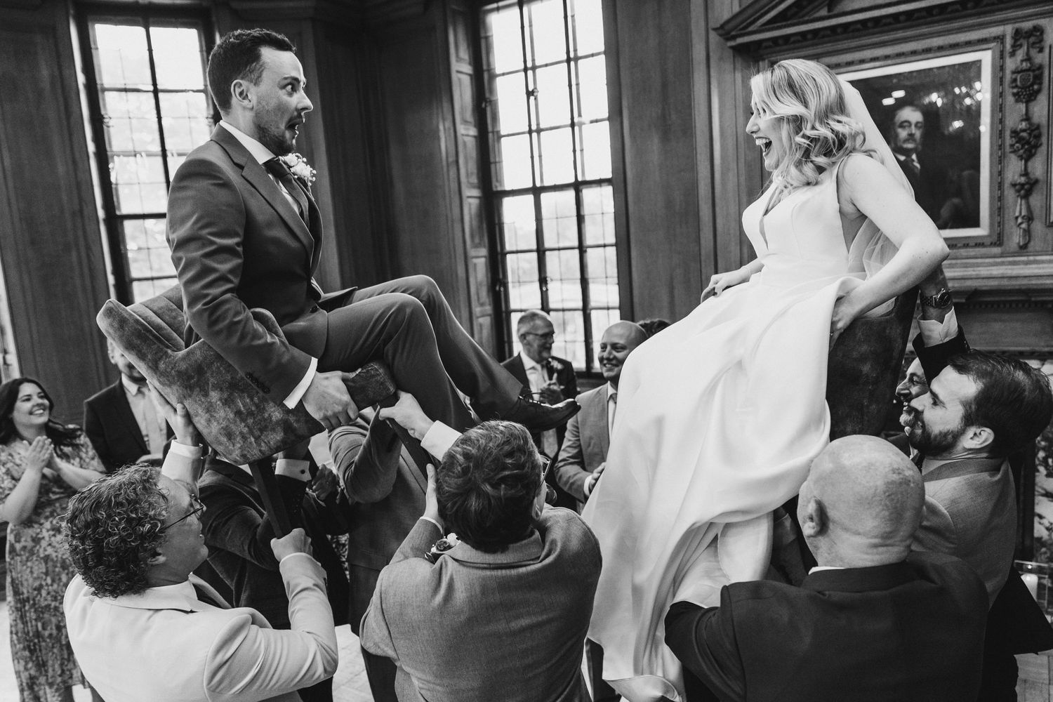 Wedding guests lift a bride and groom on chairs during a traditional Jewish hora dance celebration at an indoor venue.