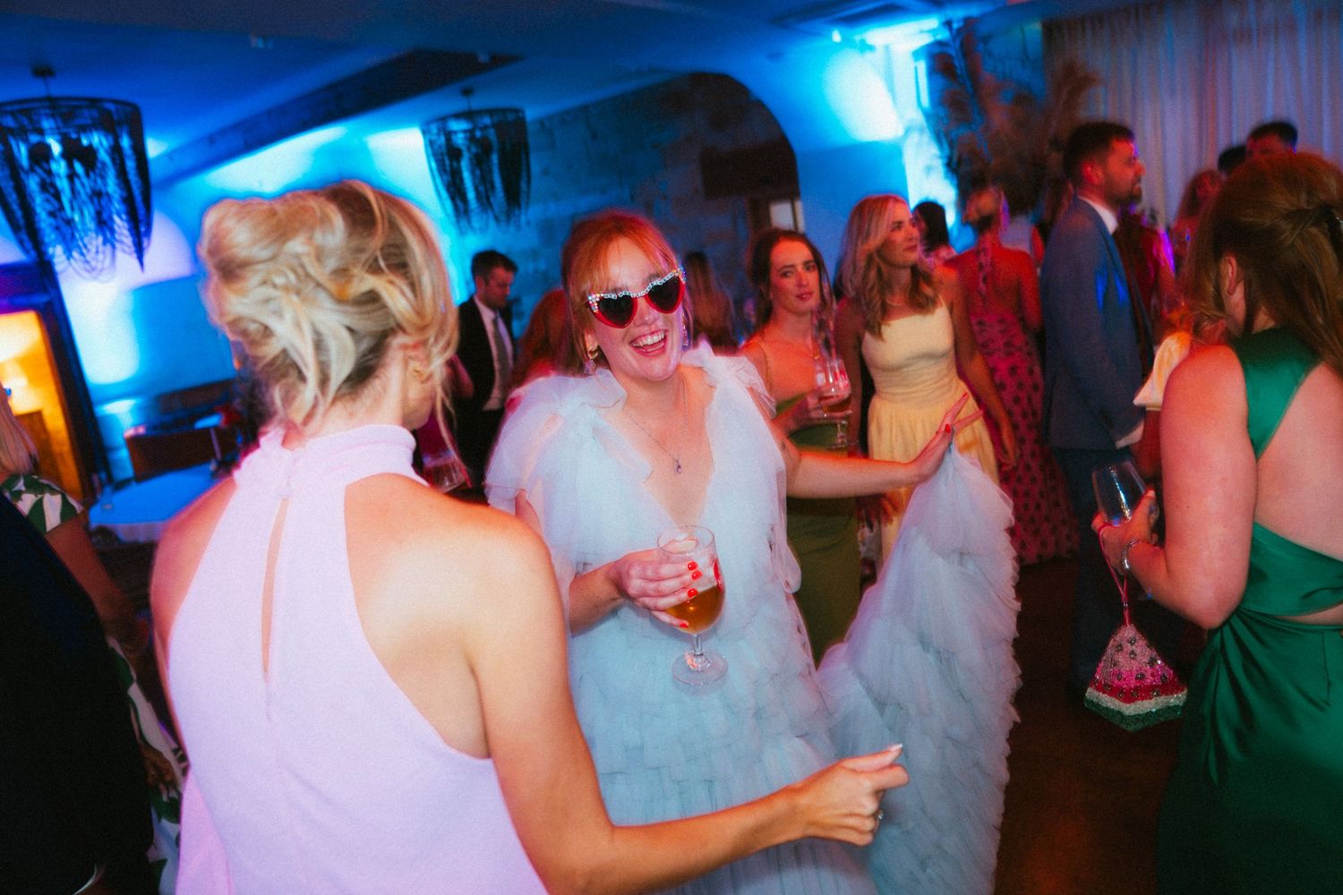 Guests in elegant dresses dance and celebrate at a wedding reception under blue lighting, with a woman in sunglasses smiling.