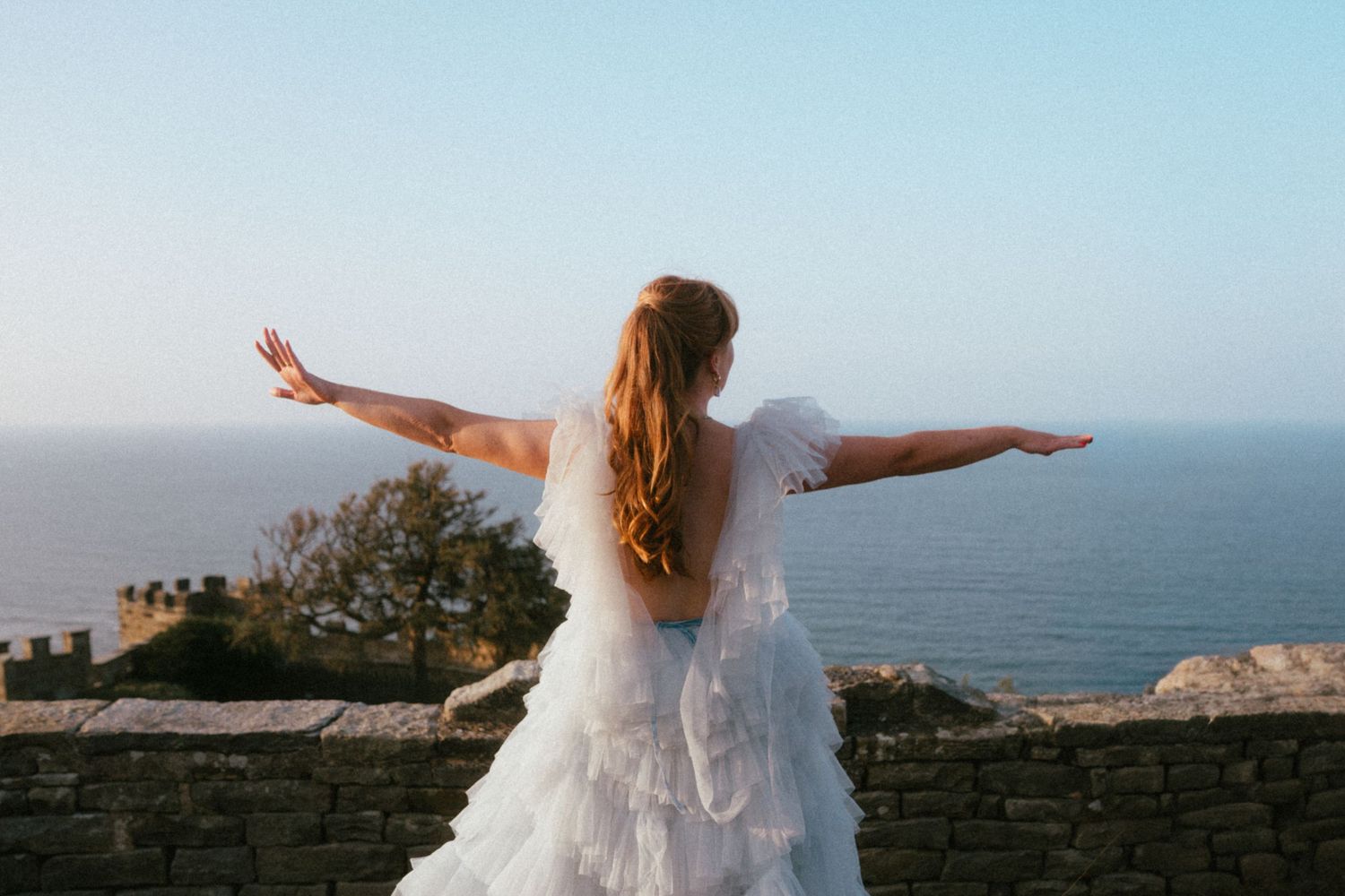 Woman in a white tulle gown with arms outstretched, seen from behind, overlooking a calm blue sea from a stone wall at sunset.