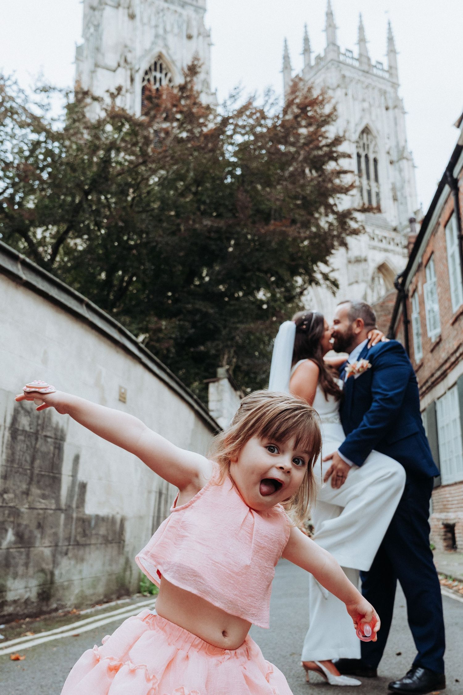 Excited flower girl in pink tutu spreads arms wide as bride and groom kiss near York Minster cathedral.