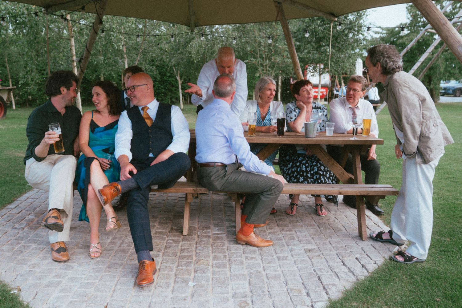 Wedding guests socializing under a rustic wooden gazebo at an outdoor reception.