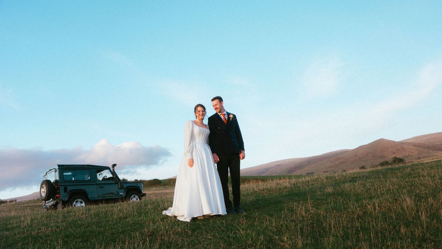 Bride in a white gown and groom in a dark suit stand on a grassy hillside beside a green 4x4 vehicle at sunset.