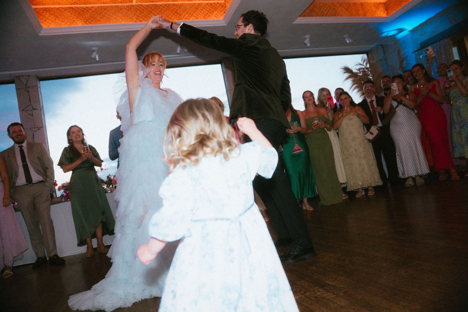 Bride in a flowing gown dances with her groom while a young flower girl twirls nearby and wedding guests watch from the dance floor.