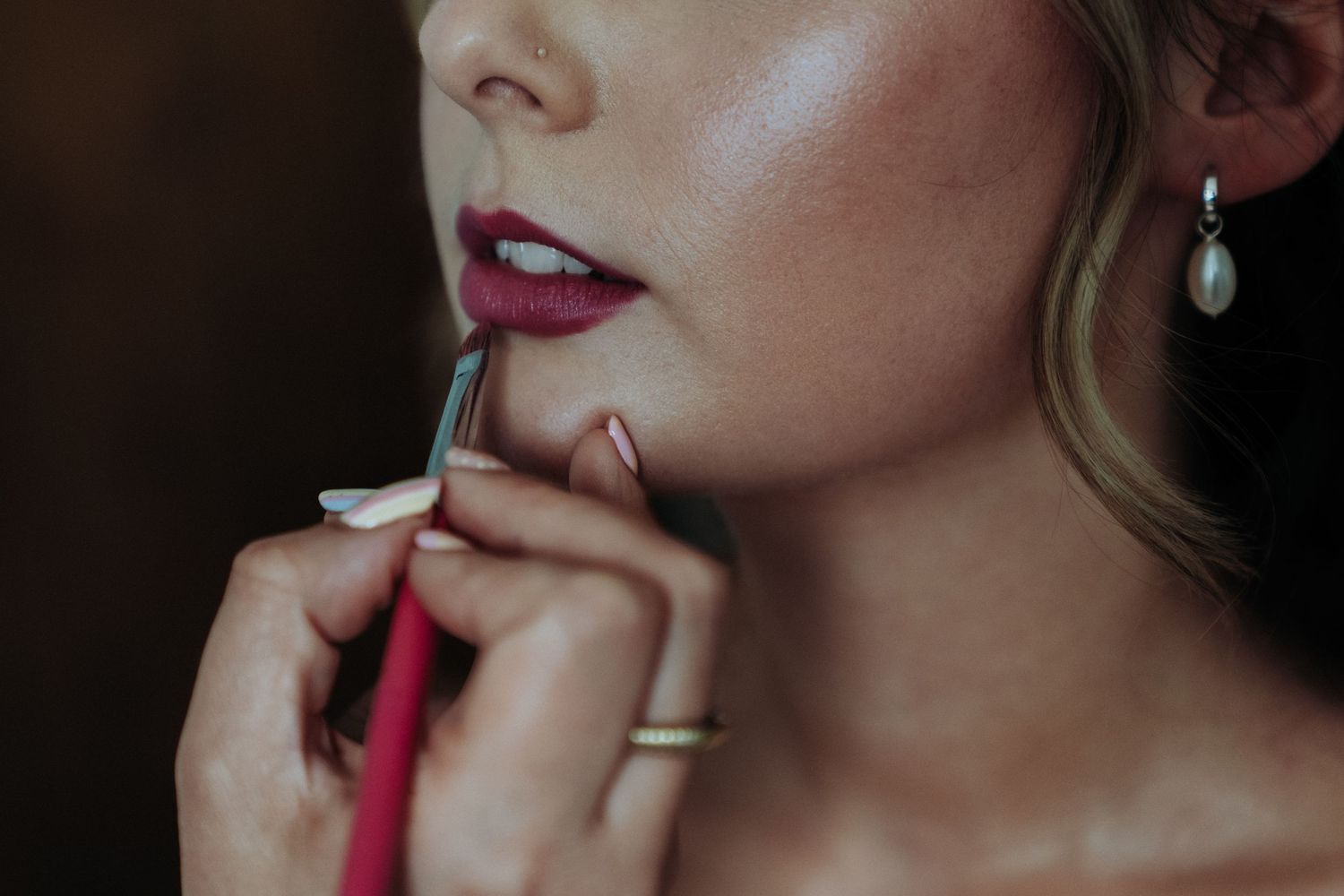 Close-up of bride having deep red lipstick applied by makeup artist with pearl earring visible.
