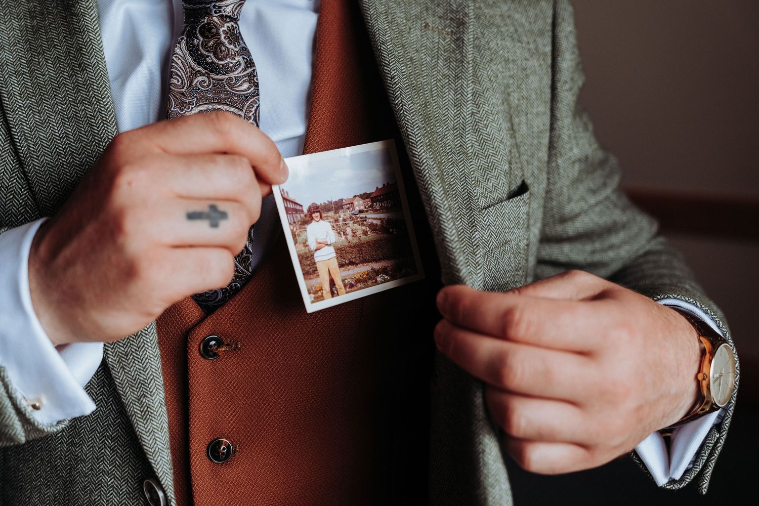 Groom in tweed suit tucking a vintage polaroid photo into his breast pocket on wedding day.