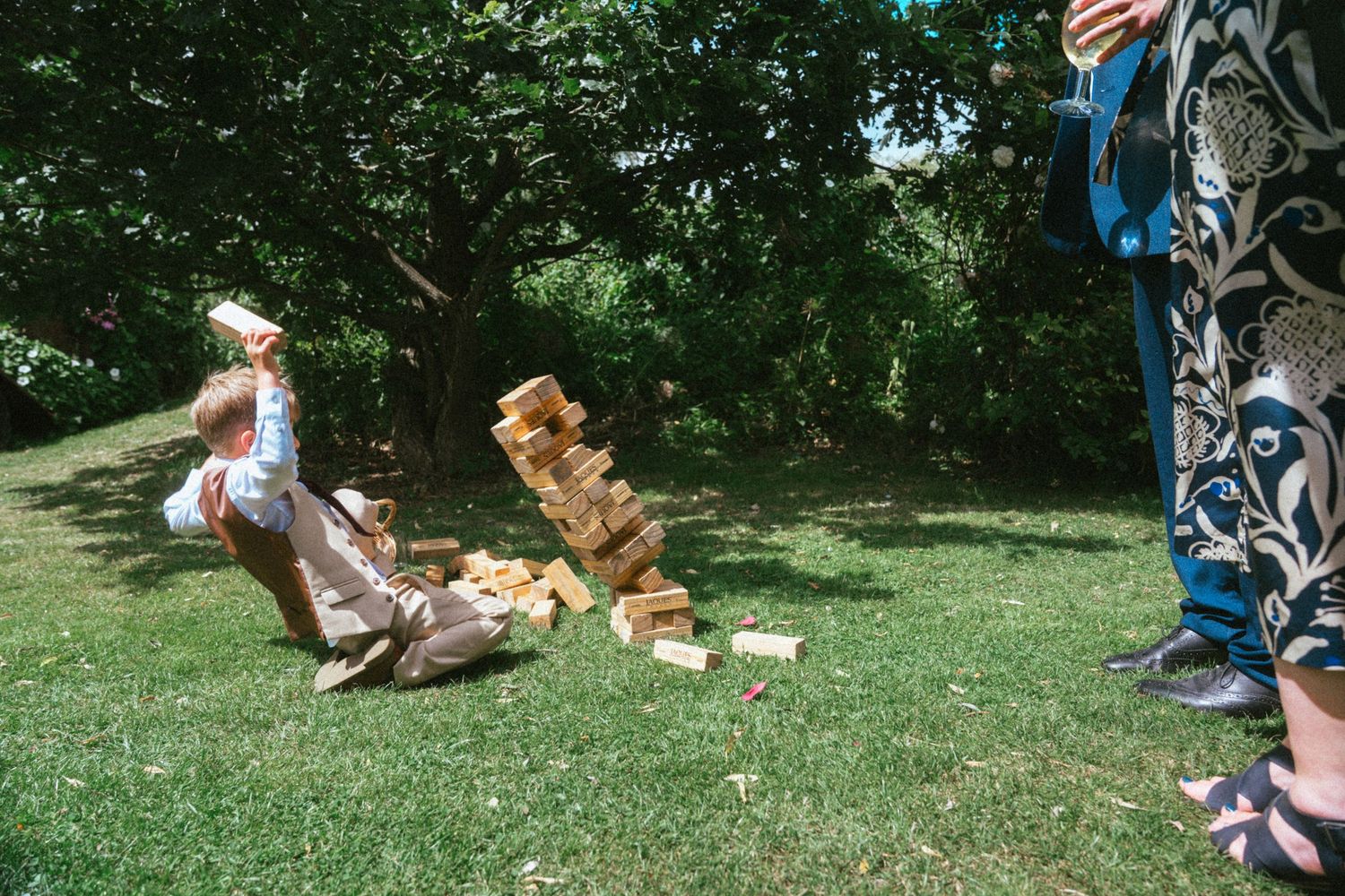 Guest falls back laughing as giant Jenga tower topples over on green lawn at outdoor wedding.