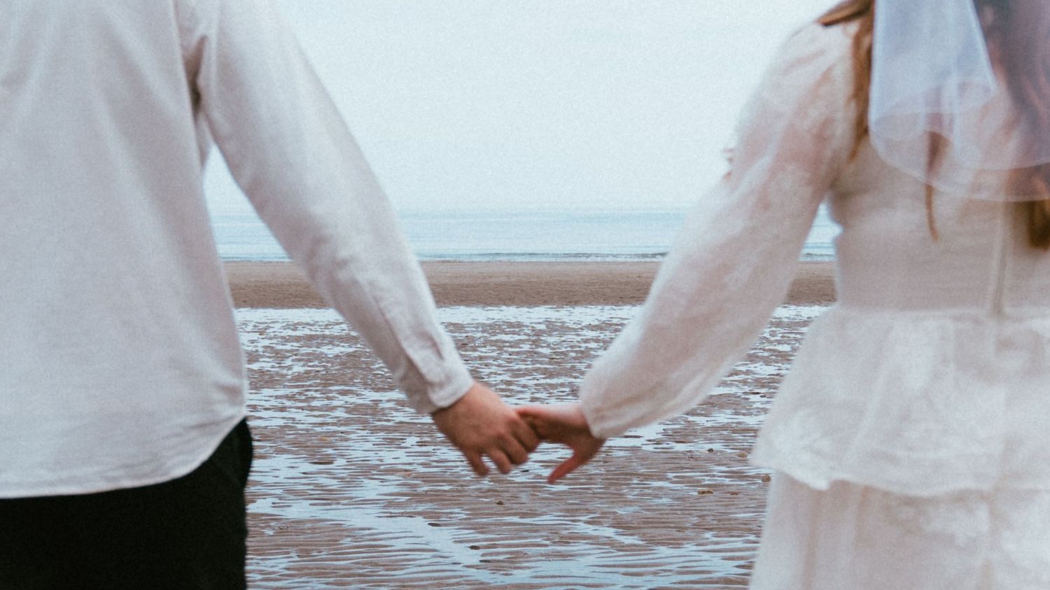 Bride in a white veil and groom in a white shirt hold hands while standing on a wet sandy beach facing the sea.