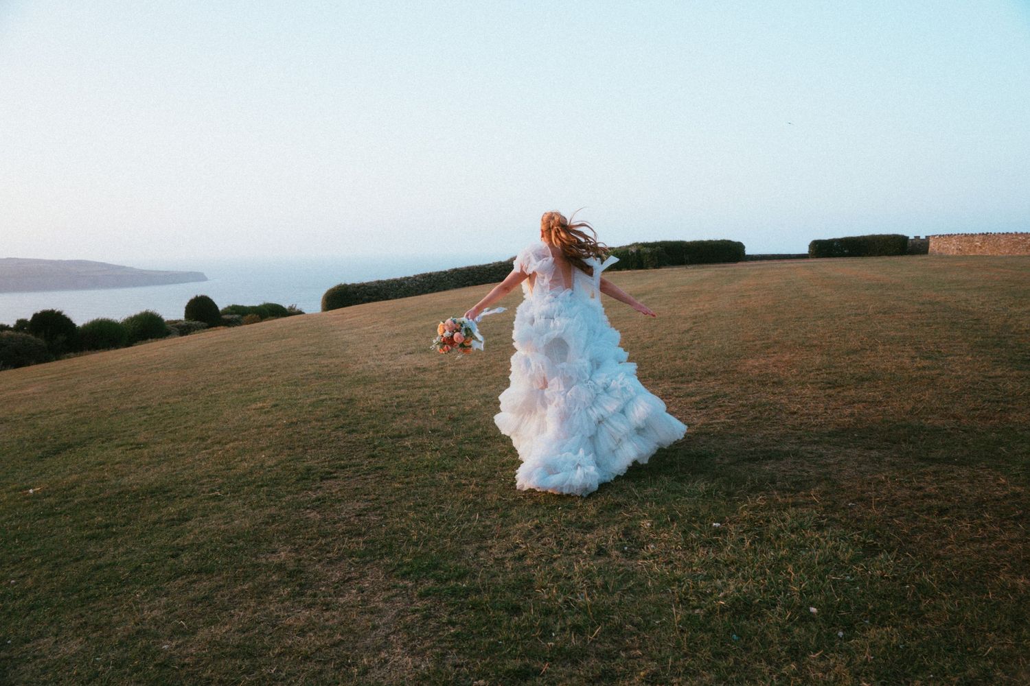 Bride in a flowing white ruffled wedding gown twirls with a bouquet on a grassy hillside overlooking the sea at sunset.