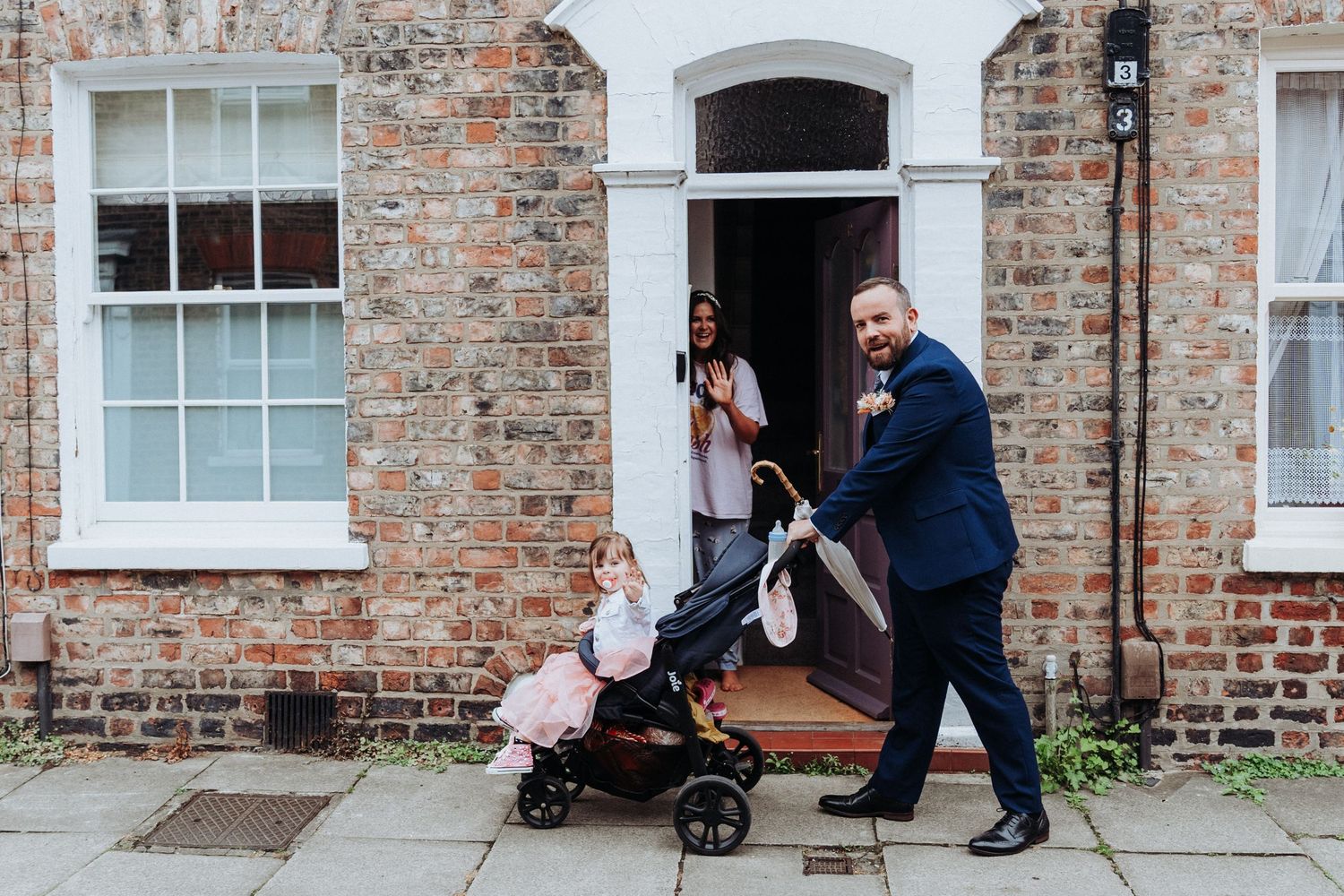 Groom in navy suit pushing a stroller outside a brick townhouse with family in doorway.